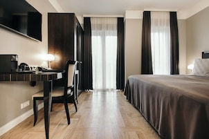 Una camera d'albergo con un letto, una scrivania con una sedia, una televisione e un'ampia finestra con tende.