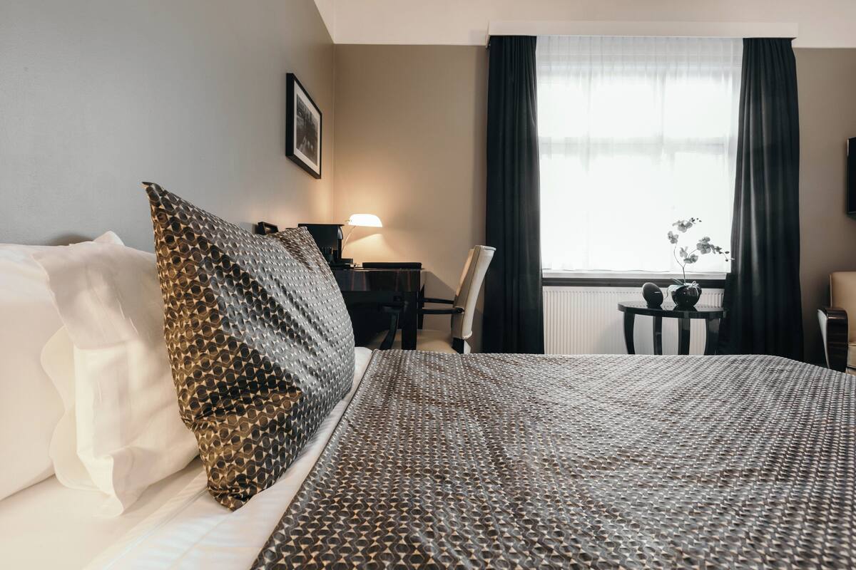 Siisti hotellihuone, jossa on siististi pedattu sänky, työpöytä lampun kanssa, tuoli ja ikkuna, jossa on verhot.