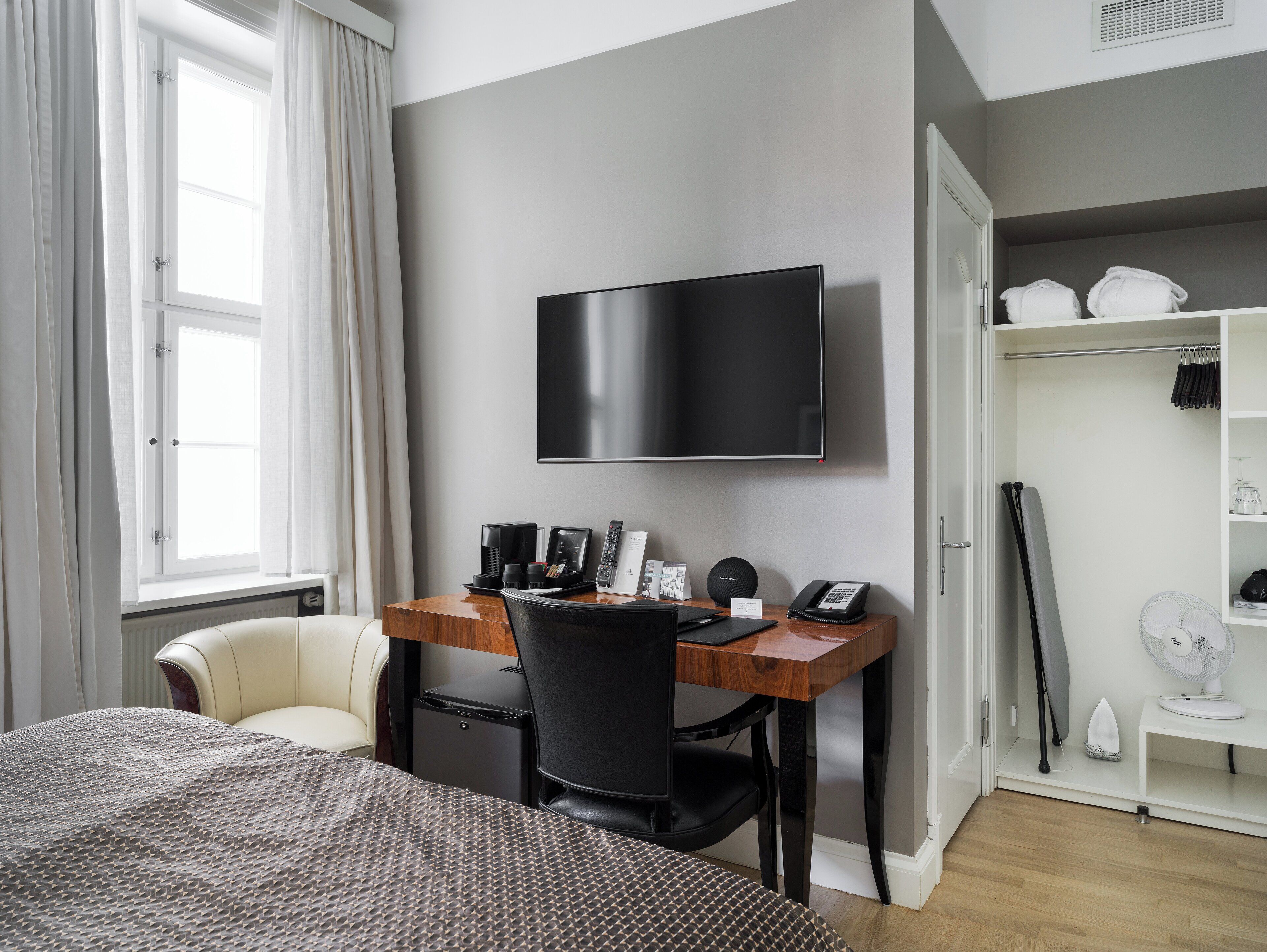 Une chambre d’hôtel comprenant un lit, un bureau avec un téléviseur à écran plat, une chaise, une petite lampe de bureau, un téléphone, une machine à café et une étagère avec des serviettes.
