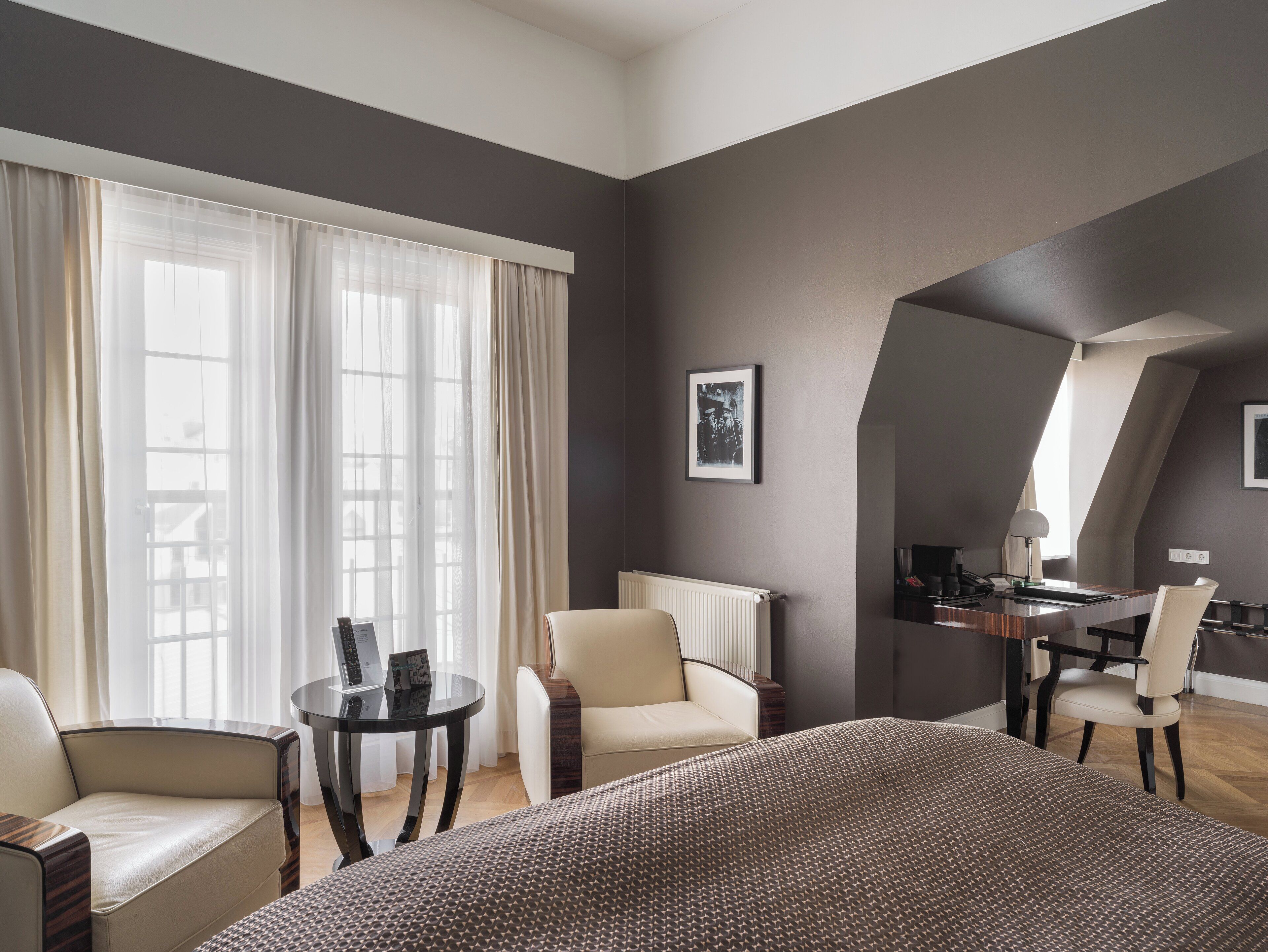 A modern hotel room with a large bed, two armchairs, a small round table, and a desk with a chair. There is a window with sheer curtains and a radiator on the wall.