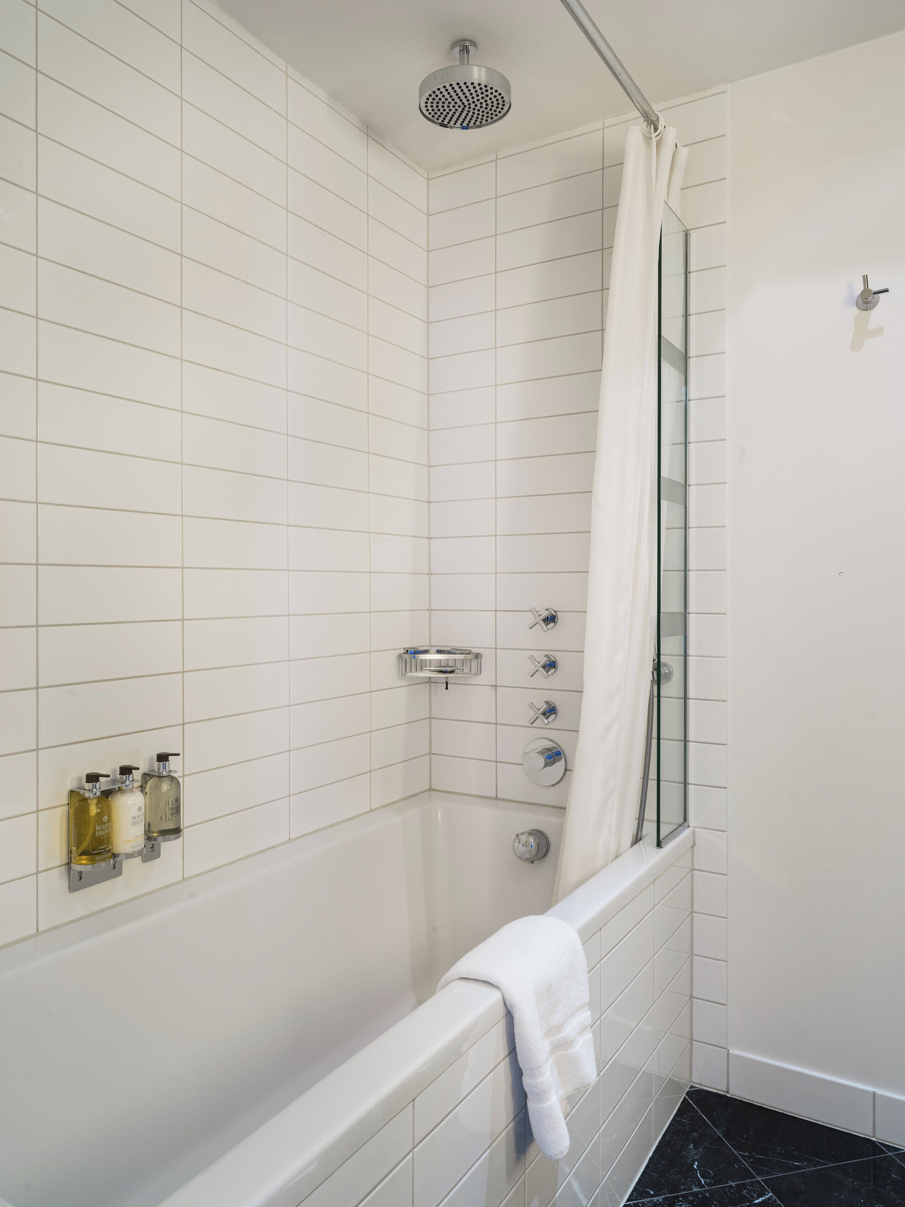 A modern bathroom with white tiled walls, a glass shower enclosure, a bathtub, and three hand soap dispensers mounted on the wall.