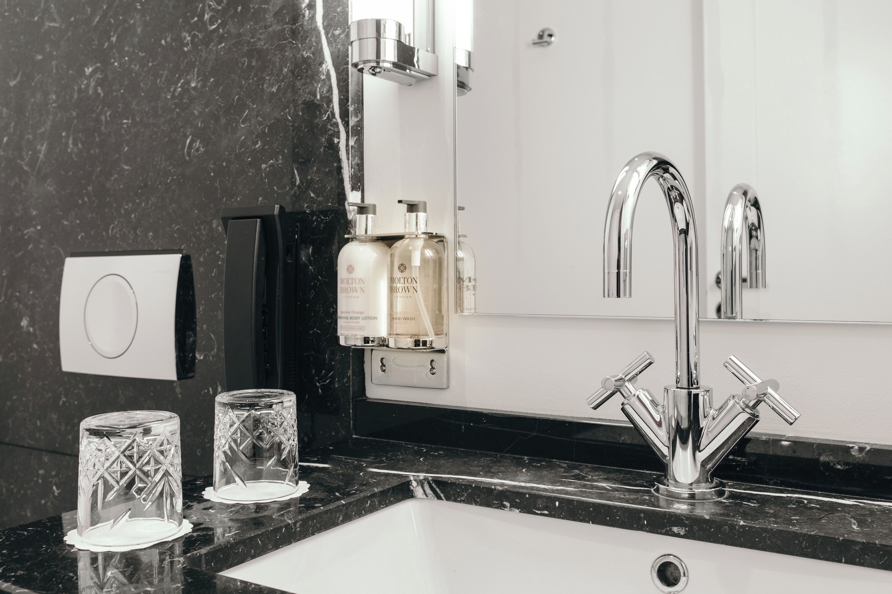 Un baño moderno con encimera de mármol negro, un lavamanos blanco y un grifo de cromo. Hay dos botellas de loción en la pared y dos vasos de vidrio transparente sobre la encimera.