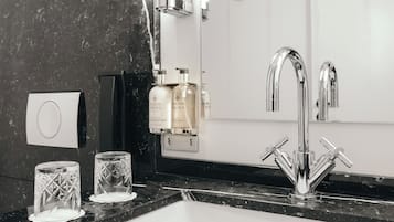 Un baño moderno con encimera de mármol negro, un lavamanos blanco y un grifo de cromo. Hay dos botellas de loción en la pared y dos vasos de vidrio transparente sobre la encimera.