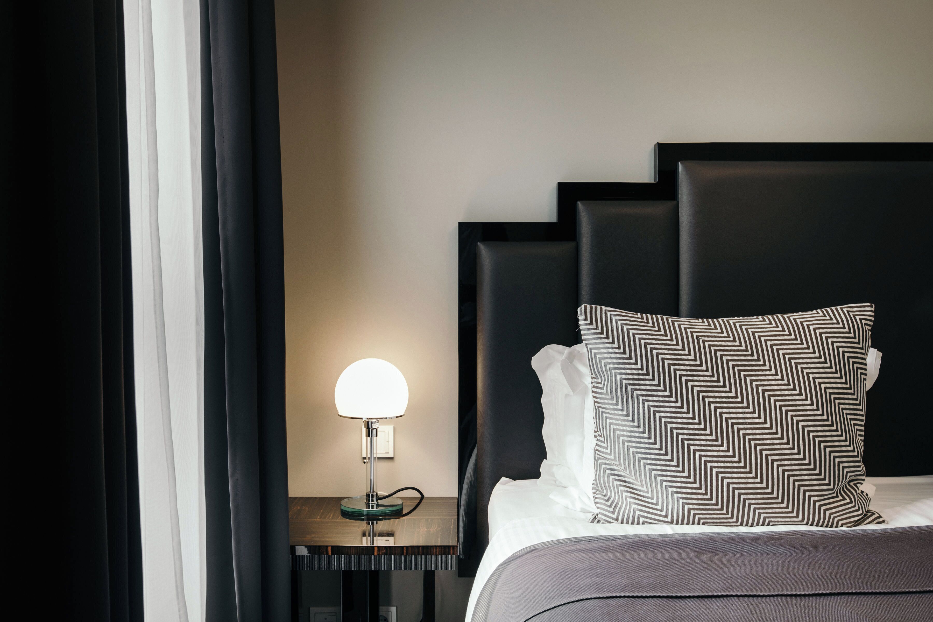Una cama bien hecha con un cojín estampado y una lámpara de noche sobre una mesita de madera oscura.