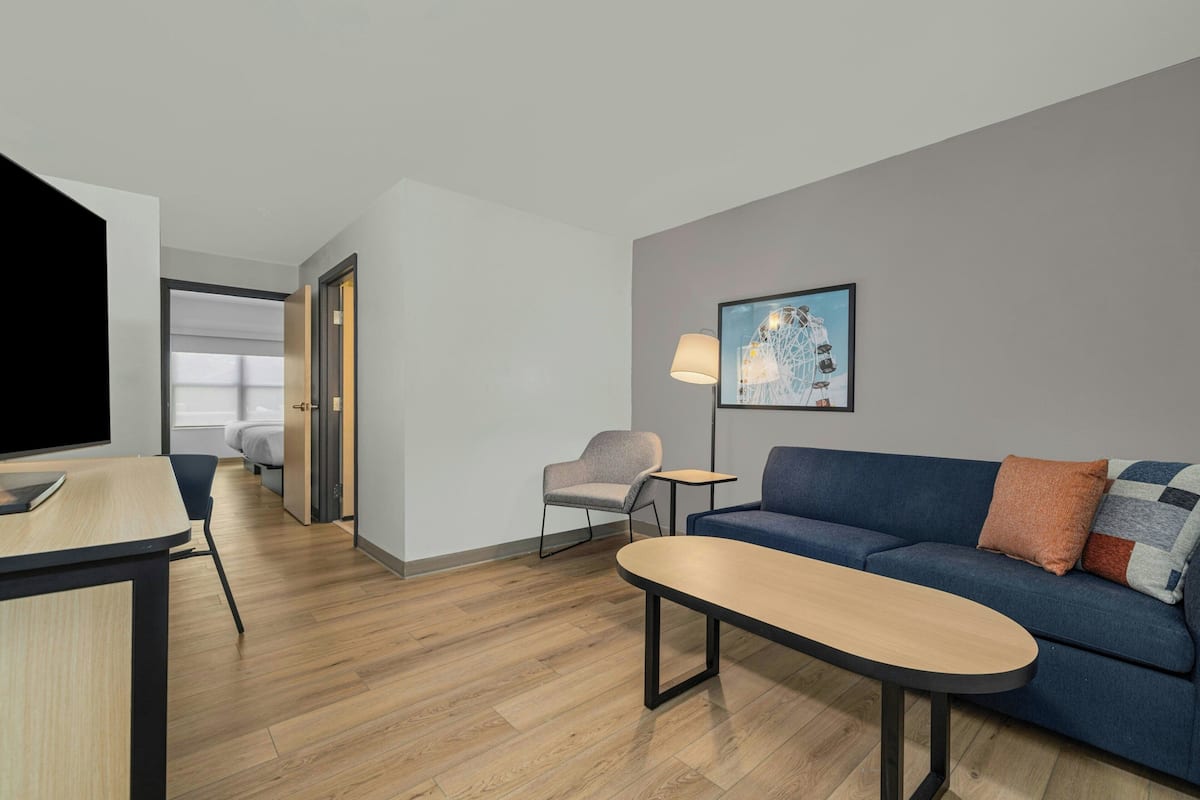 A modern hotel room with a wooden floor, a sofa, a small table, a chair, a TV, and a framed picture on the wall.