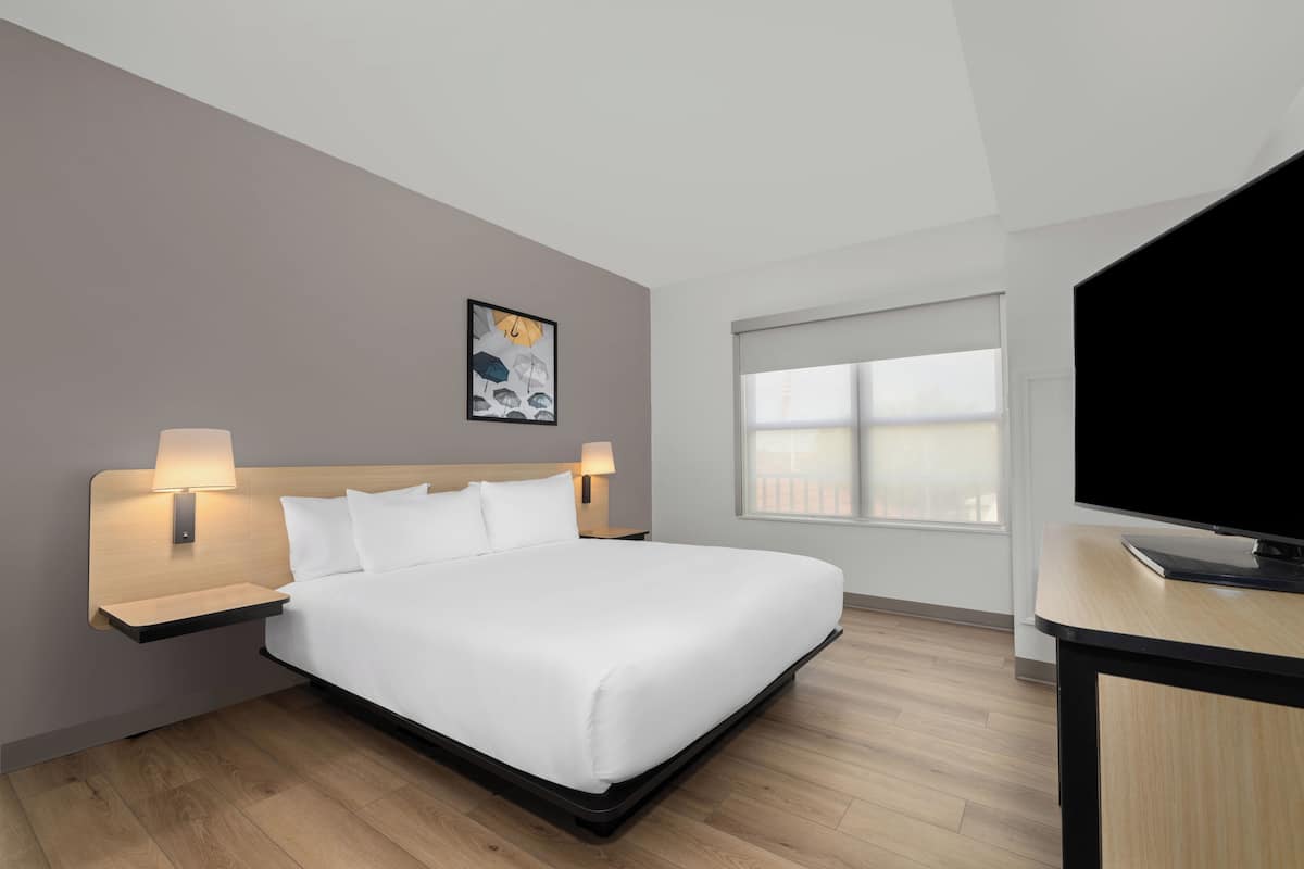 A modern hotel room with a large bed, bedside lamps, a flat-screen TV, and a window with blinds.