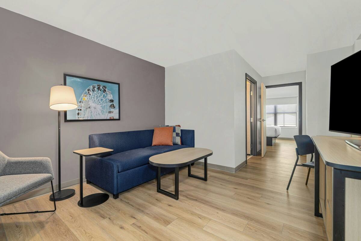 A modern hotel room with a sofa, a small table, a floor lamp, a TV, and a painting of a Ferris wheel.
