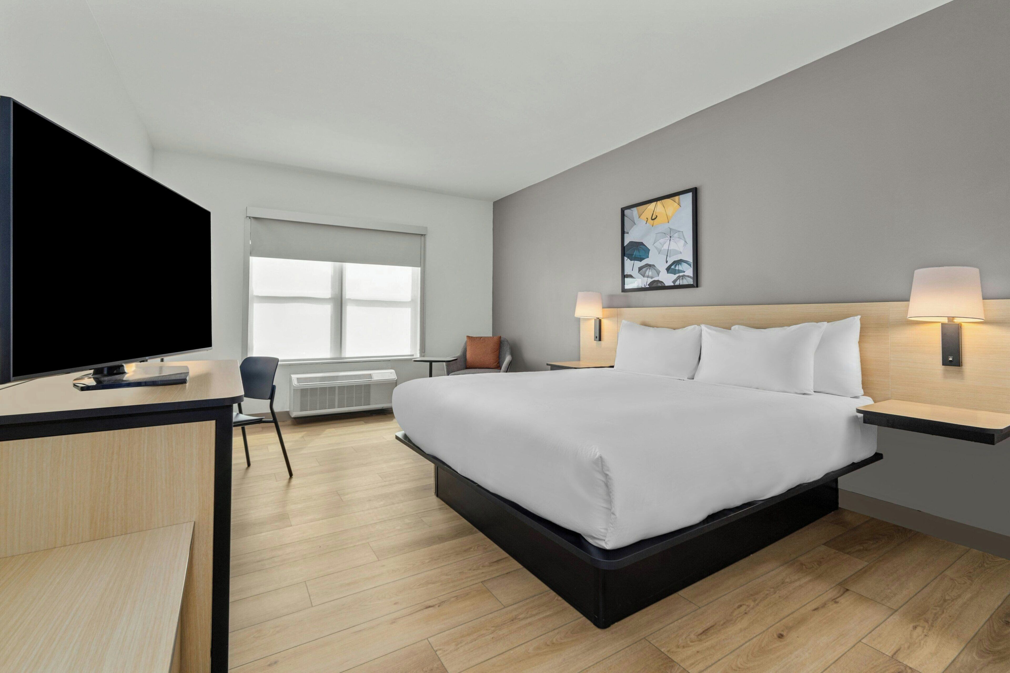 A modern hotel room with a bed, bedside lamps, a desk with a flat-screen TV, and a window.