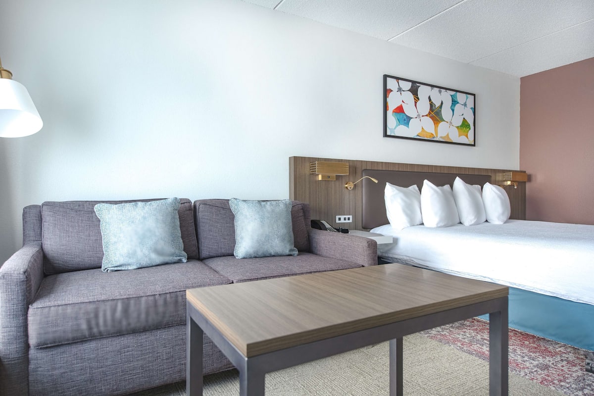 Habitación de hotel con un sofá gris, una mesa de centro de madera, una cama con ropa de cama blanca y un cuadro abstracto colorido en la pared.