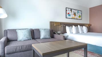 A hotel room with a grey sofa, a wooden coffee table, a bed with white bedding, and a colorful abstract painting on the wall.