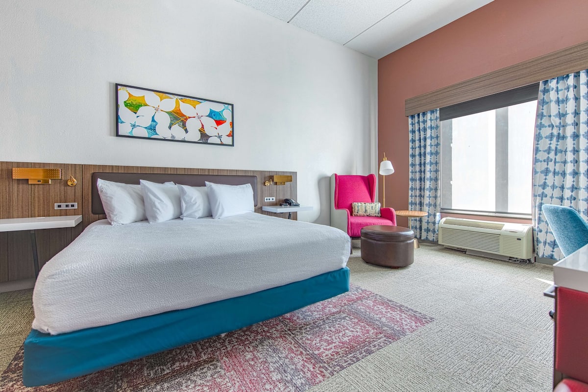 Habitación de hotel con una cama grande, una mesita de noche, una silla, un ventanal con cortinas y una obra de arte en la pared.