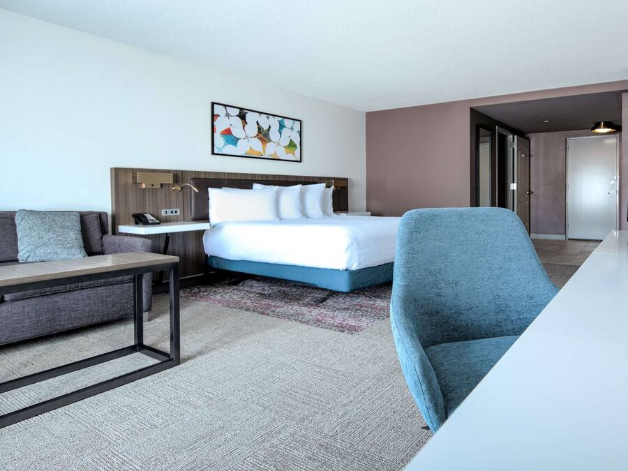 A modern hotel room with a bed, a desk, a chair, and a painting on the wall.