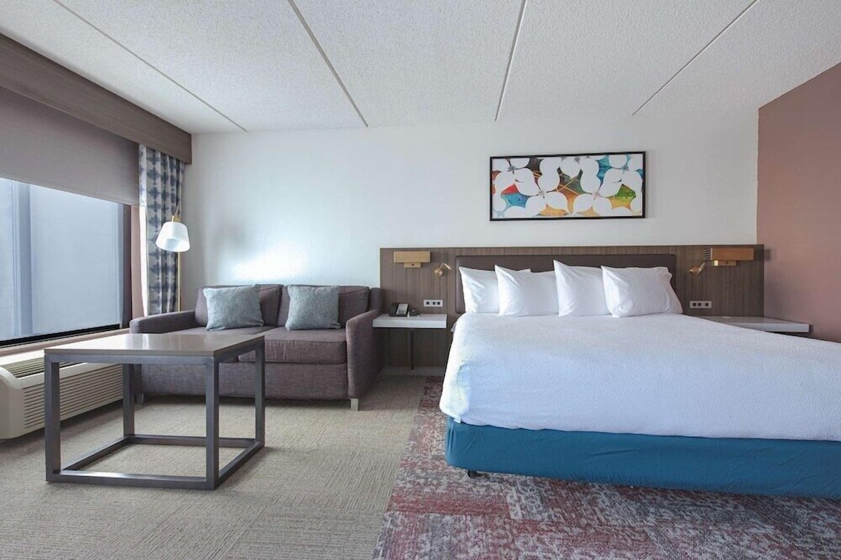 Un cuarto de hotel con cama, sofá, mesa, lámpara y un cuadro en la pared.