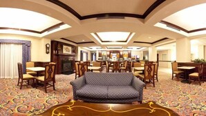 Eine Hotellobby mit einem gemusterten Teppich, einem zentralen Sofa und einem Essbereich mit Holztischen und -stühlen.