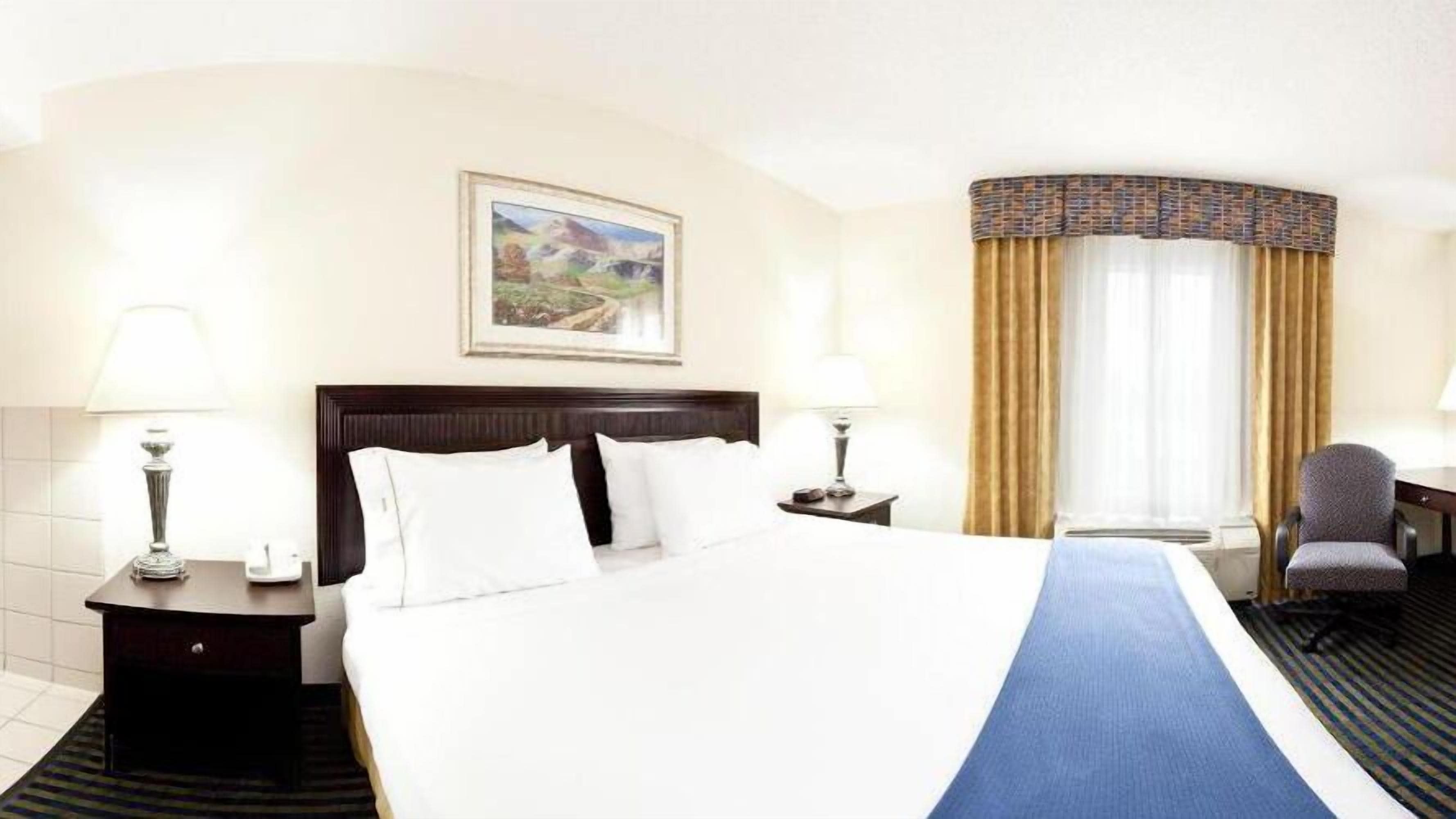 A hotel room with a large bed, two bedside tables with lamps, a desk with a chair, a window with curtains, and a framed picture on the wall.
