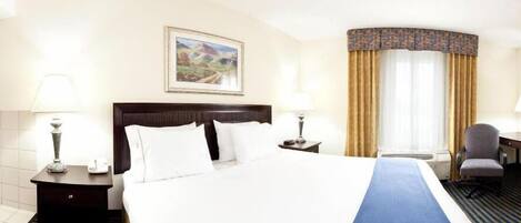 A hotel room with a large bed, two bedside tables with lamps, a desk with a chair, a window with curtains, and a framed picture on the wall.