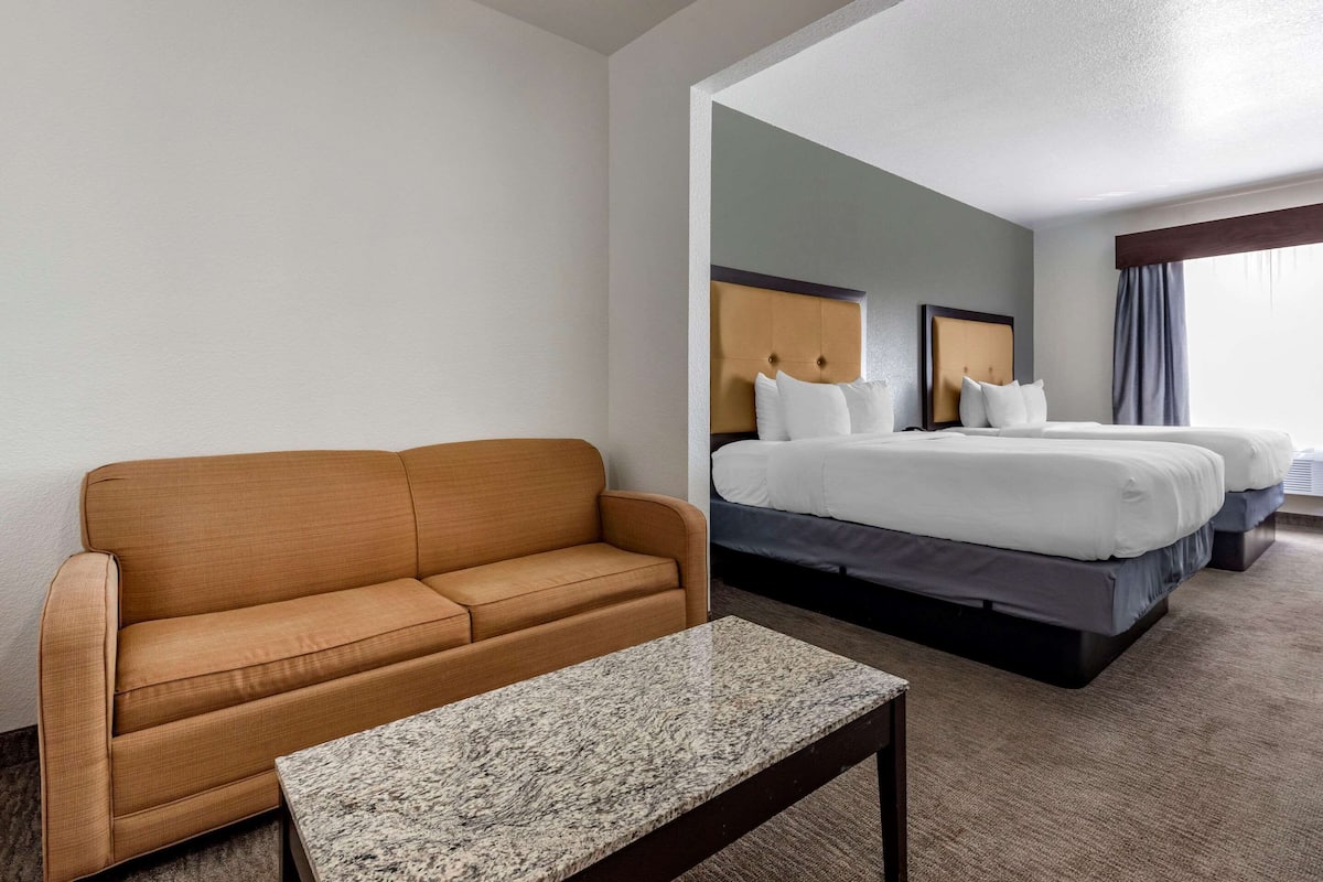 A hotel room with a sofa, a bed with two pillows, a marble table, and a large window with curtains.