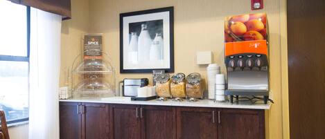 Un mostrador de desayuno de hotel con cafetera, dispensador de zumo y varios recipientes de comida.