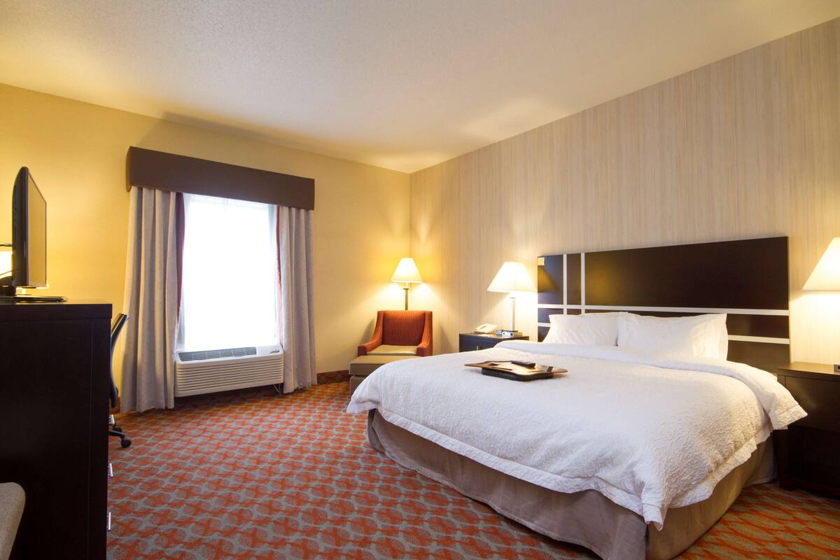 Une chambre d’hôtel avec un grand lit, deux tables de chevet avec des lampes, un bureau avec une chaise, une fenêtre avec des rideaux et un radiateur.