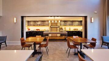 A modern dining area with wooden tables and chairs, a kitchen in the background, and pendant lighting.