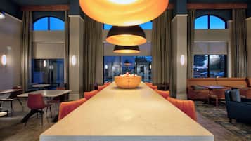A long communal dining table with a central bowl, surrounded by orange chairs, in a modern hotel lobby with large windows and pendant lighting.