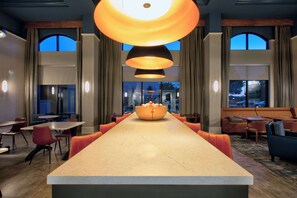 A long communal dining table with a central bowl, surrounded by orange chairs, in a modern hotel lobby with large windows and pendant lighting. - Hampton Inn & Suites Spartanburg-I-26-Westgate Mall (Spartanburg)