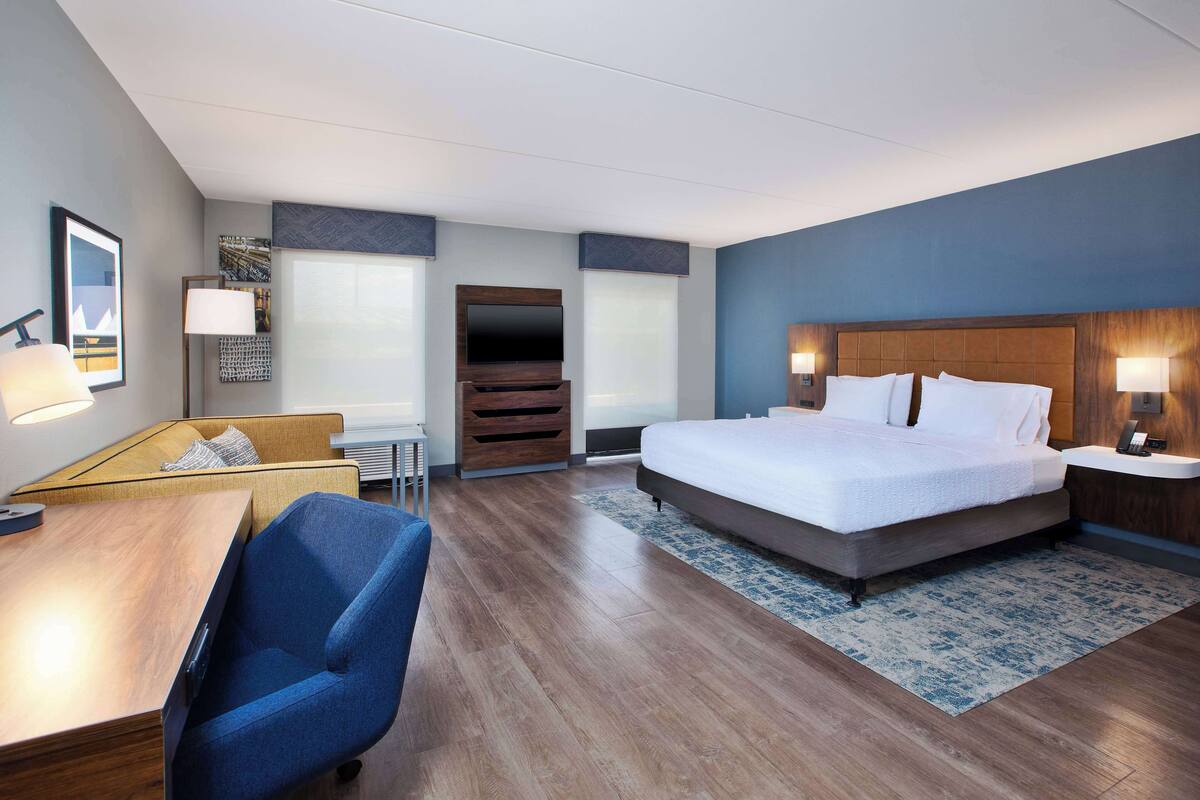 A modern hotel room with a large bed, a desk, a chair, a TV, and a small seating area.