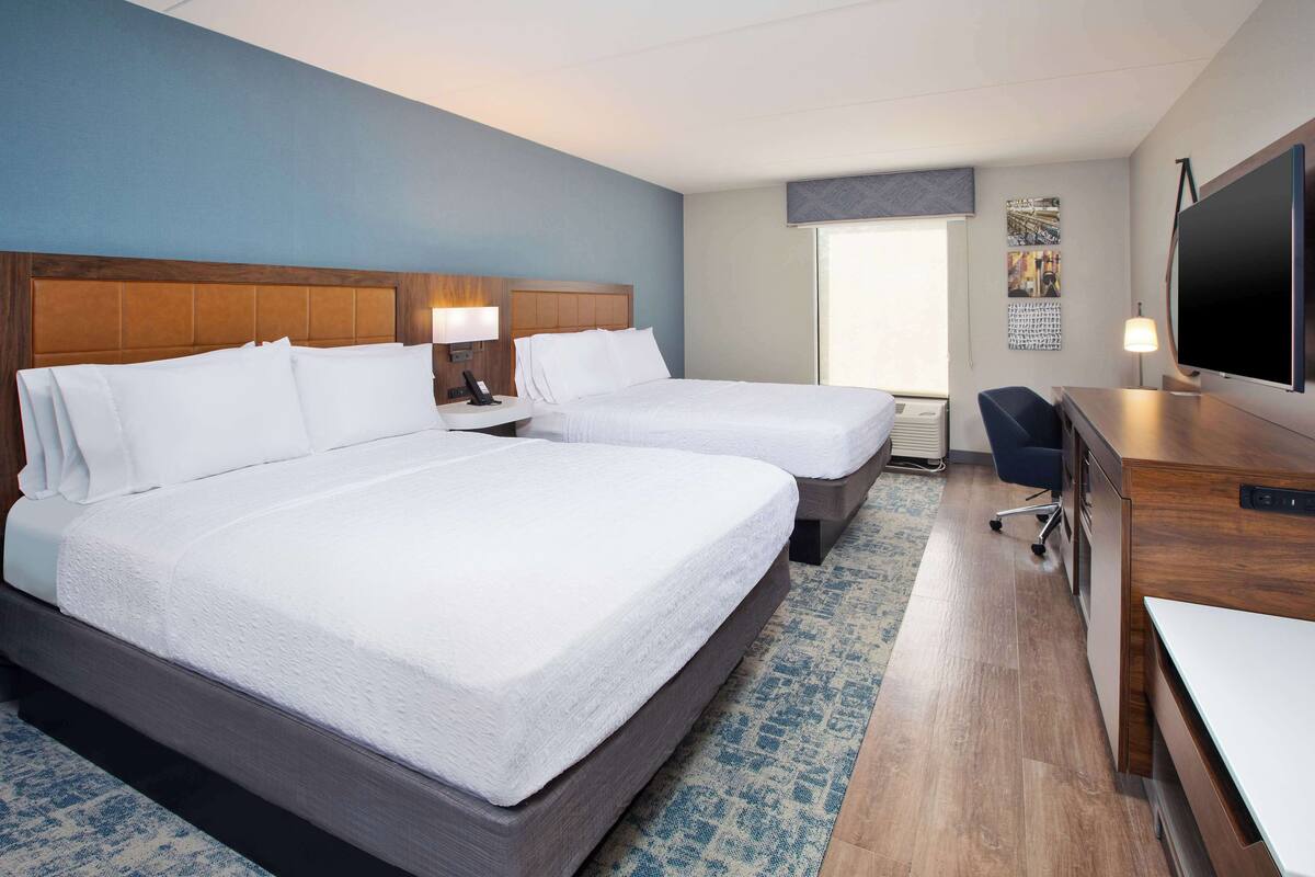 A hotel room with two beds, a desk, a TV, and a window.