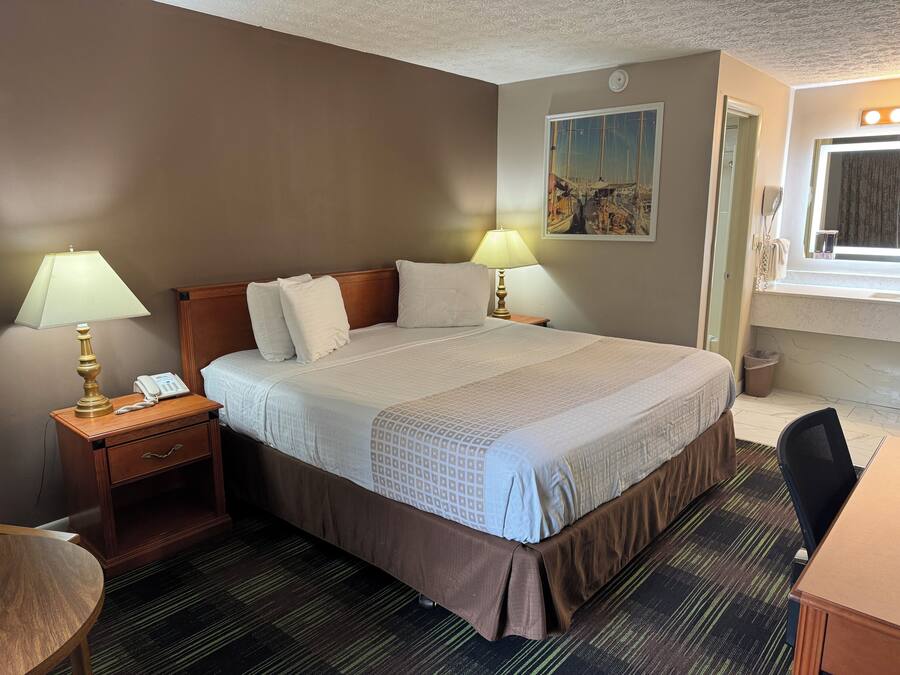 A hotel room with a large bed, two bedside lamps, a nightstand with a phone, a desk with a chair, and a painting on the wall.