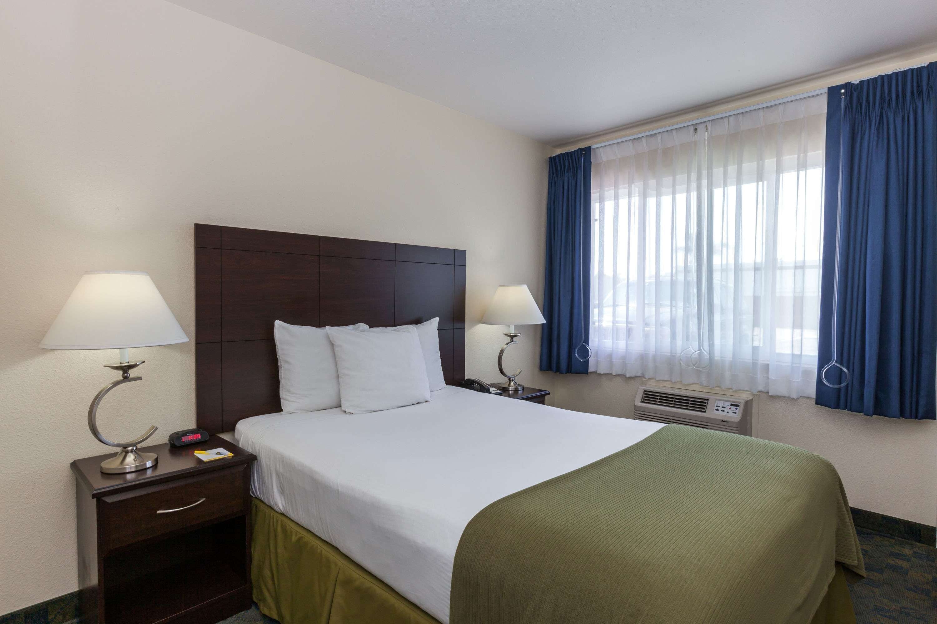Habitación de hotel con cama, dos mesitas de noche con lámparas, ventana con cortinas azules y aire acondicionado mural.