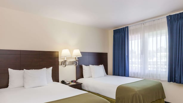 Une chambre d’hôtel avec deux lits, une table de chevet avec un téléphone, des lampes fixées au mur et une fenêtre avec des rideaux bleus.