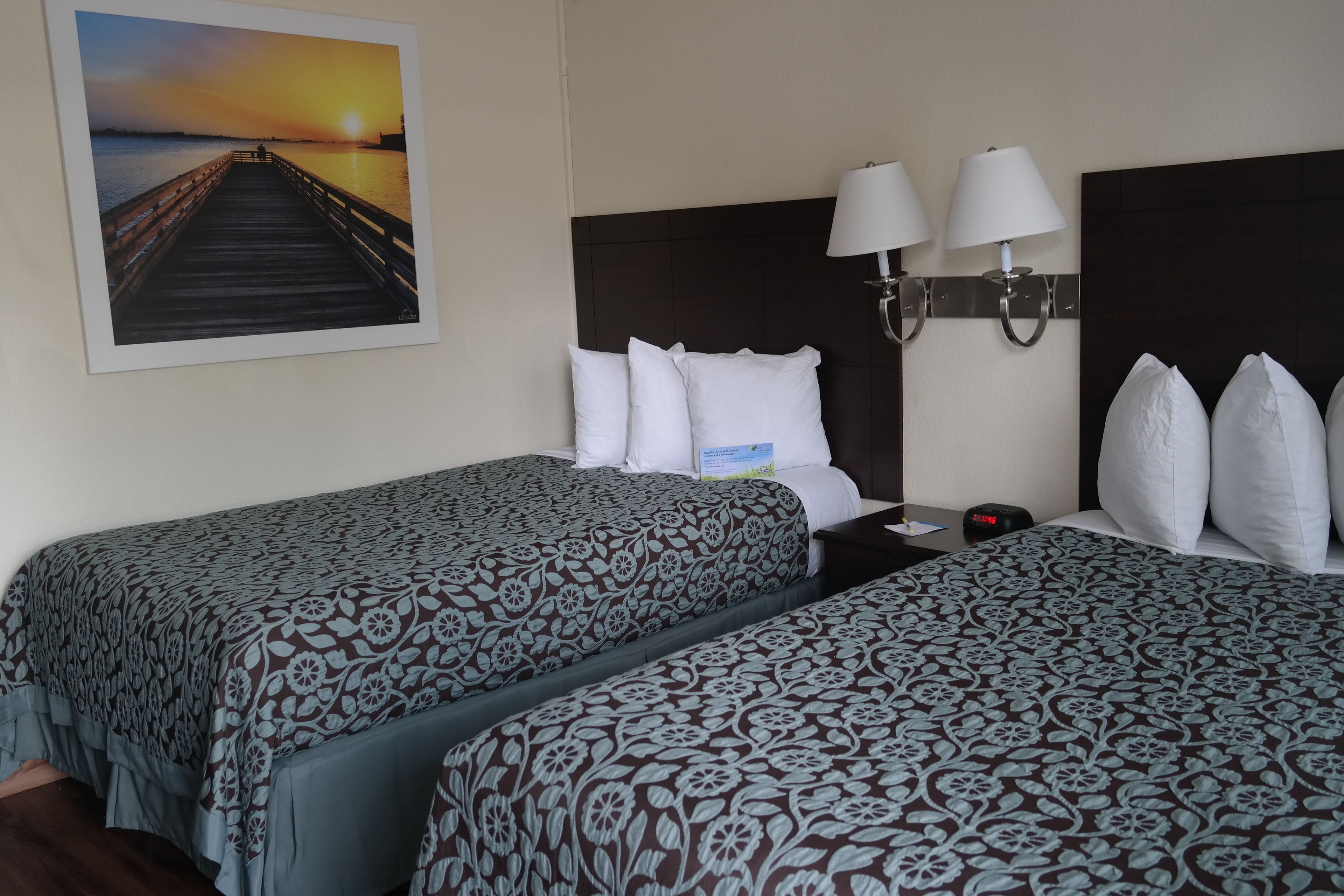 Une chambre d’hôtel avec deux lits, une table de chevet avec une lampe, une lampe fixée au mur et un tableau représentant un coucher de soleil sur une jetée.