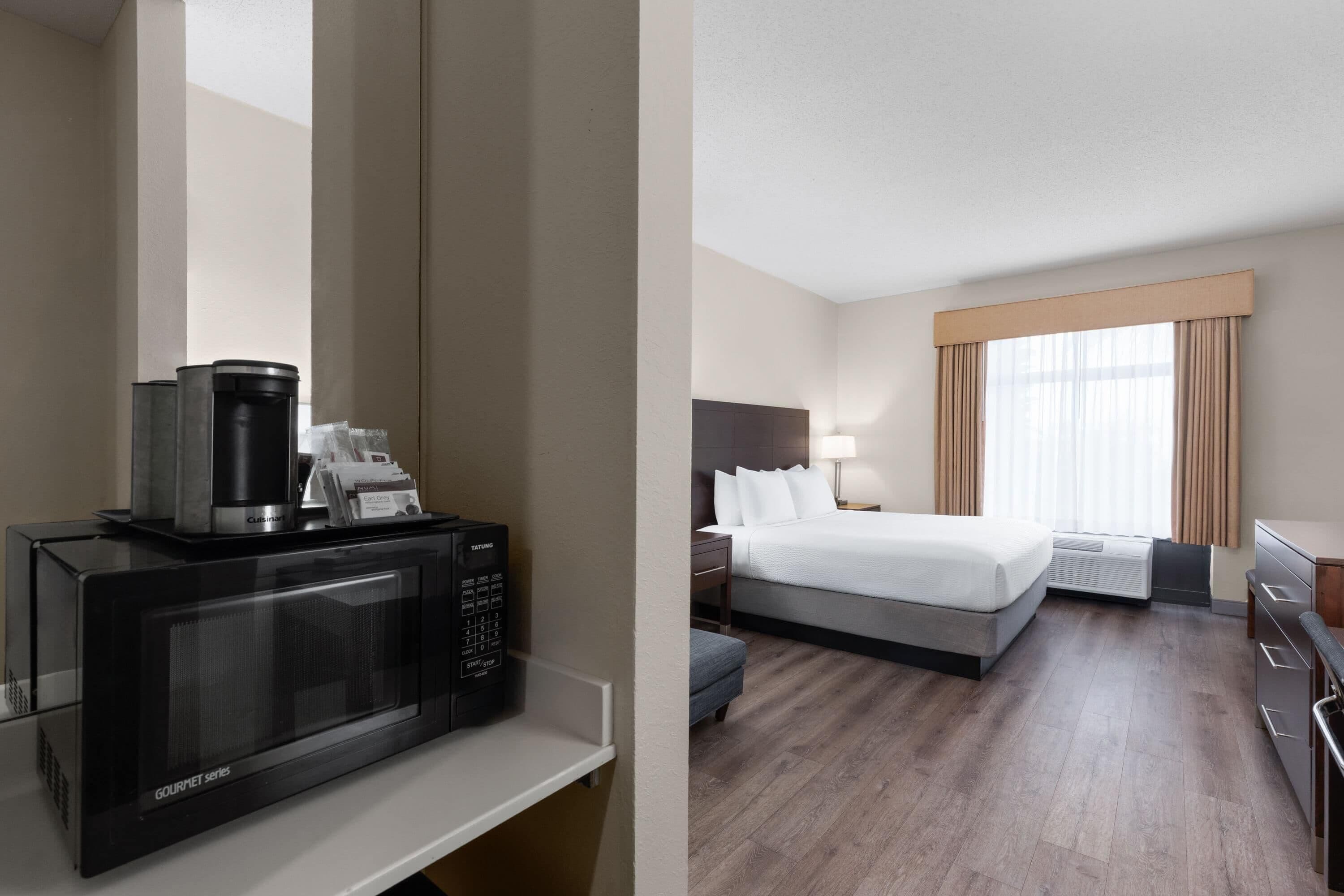 A hotel room with a microwave, a bed, a nightstand, a lamp, and a window with curtains.