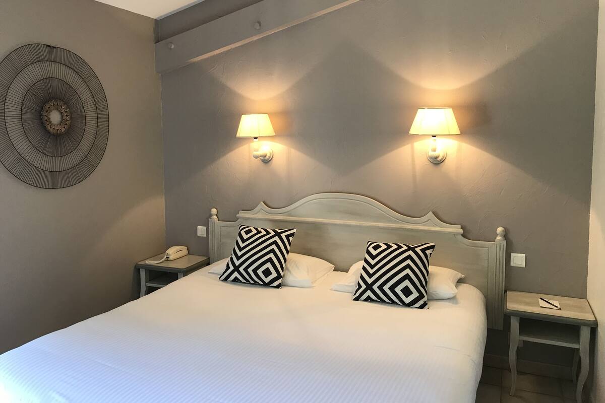 A neatly made bed with two patterned pillows, two bedside lamps, a wall clock, and a decorative wall piece.