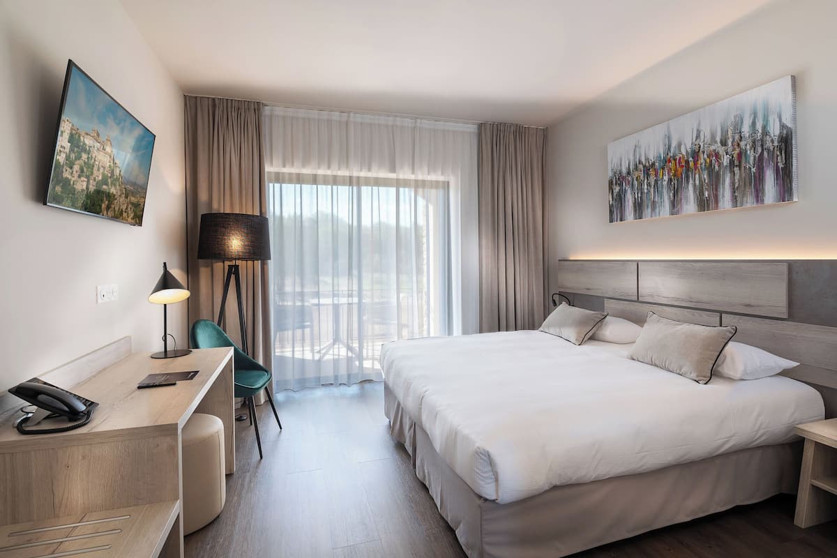 A modern hotel room with a large bed, a desk with a phone, a chair, a lamp, and a painting on the wall.