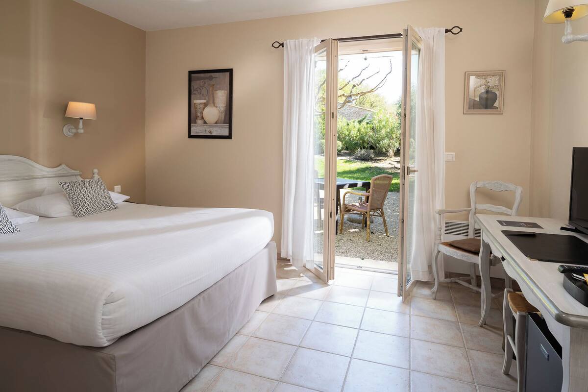 A hotel room with a bed, a desk, a chair, and a view of the garden through an open door.