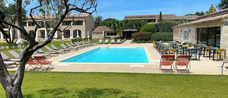 Zona de piscina con sillas de descanso y mesas, rodeada de edificios de piedra con tejados de tejas.