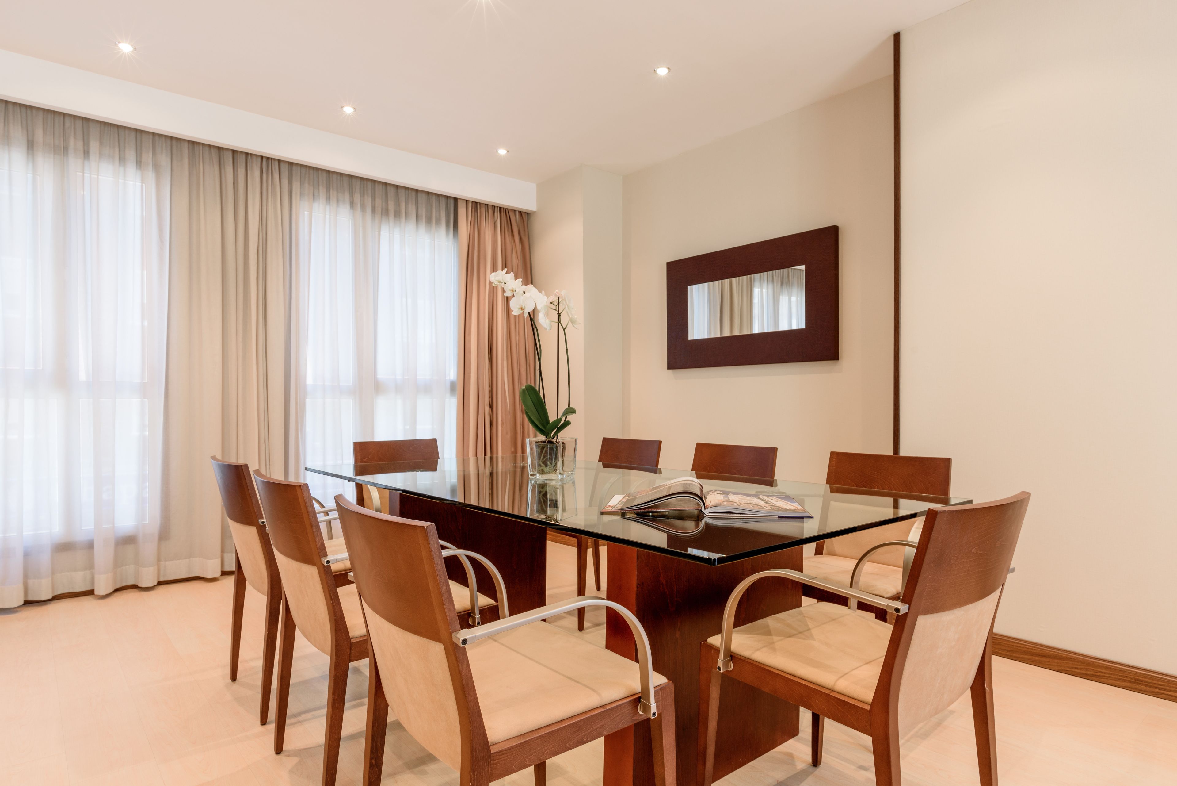A conference room with a glass table, six wooden chairs, a vase with white flowers, and a mirror on the wall.