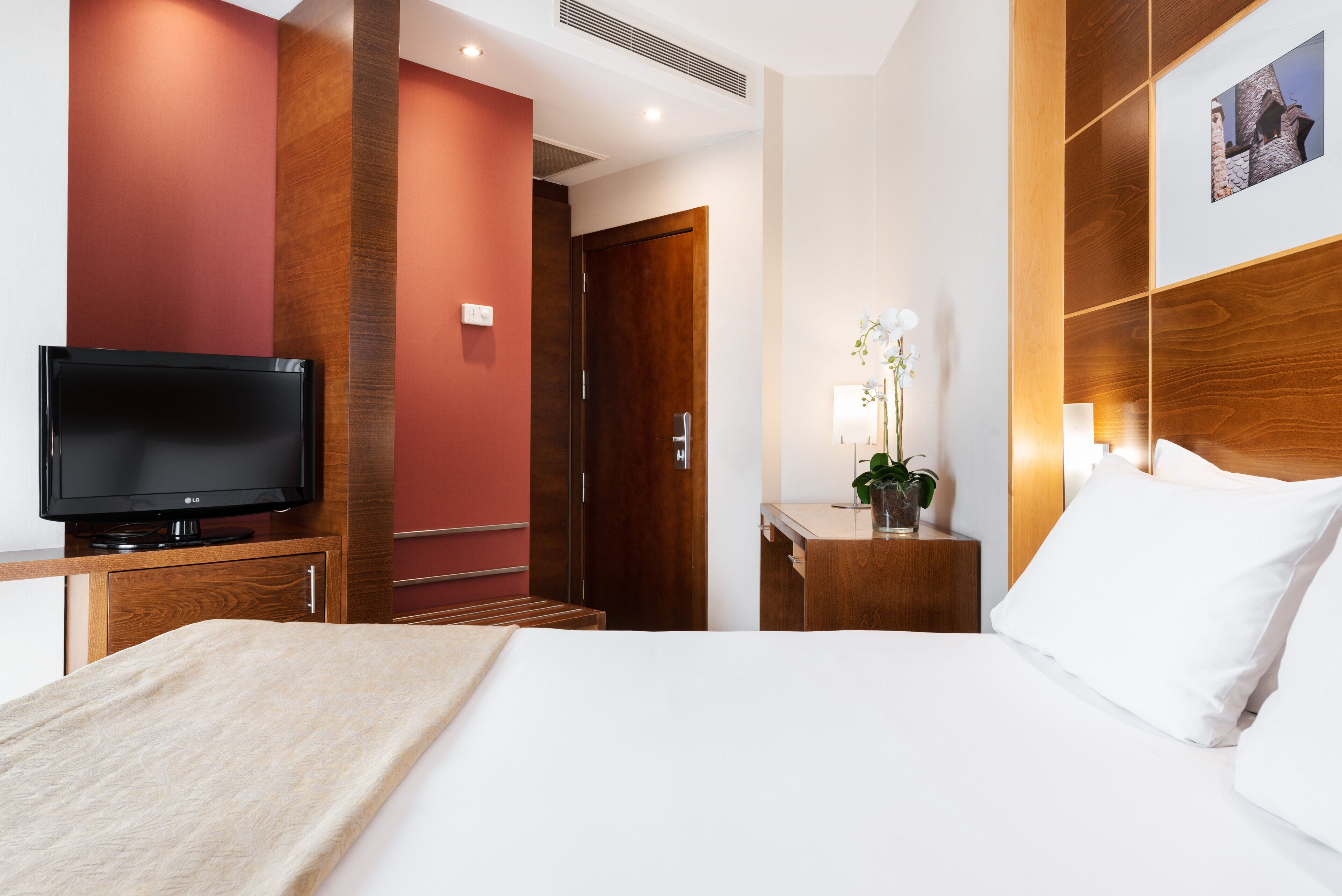 Une chambre d’hôtel avec un lit, une télévision, une tête de lit en bois, une table de chevet avec une lampe et un tableau au mur.