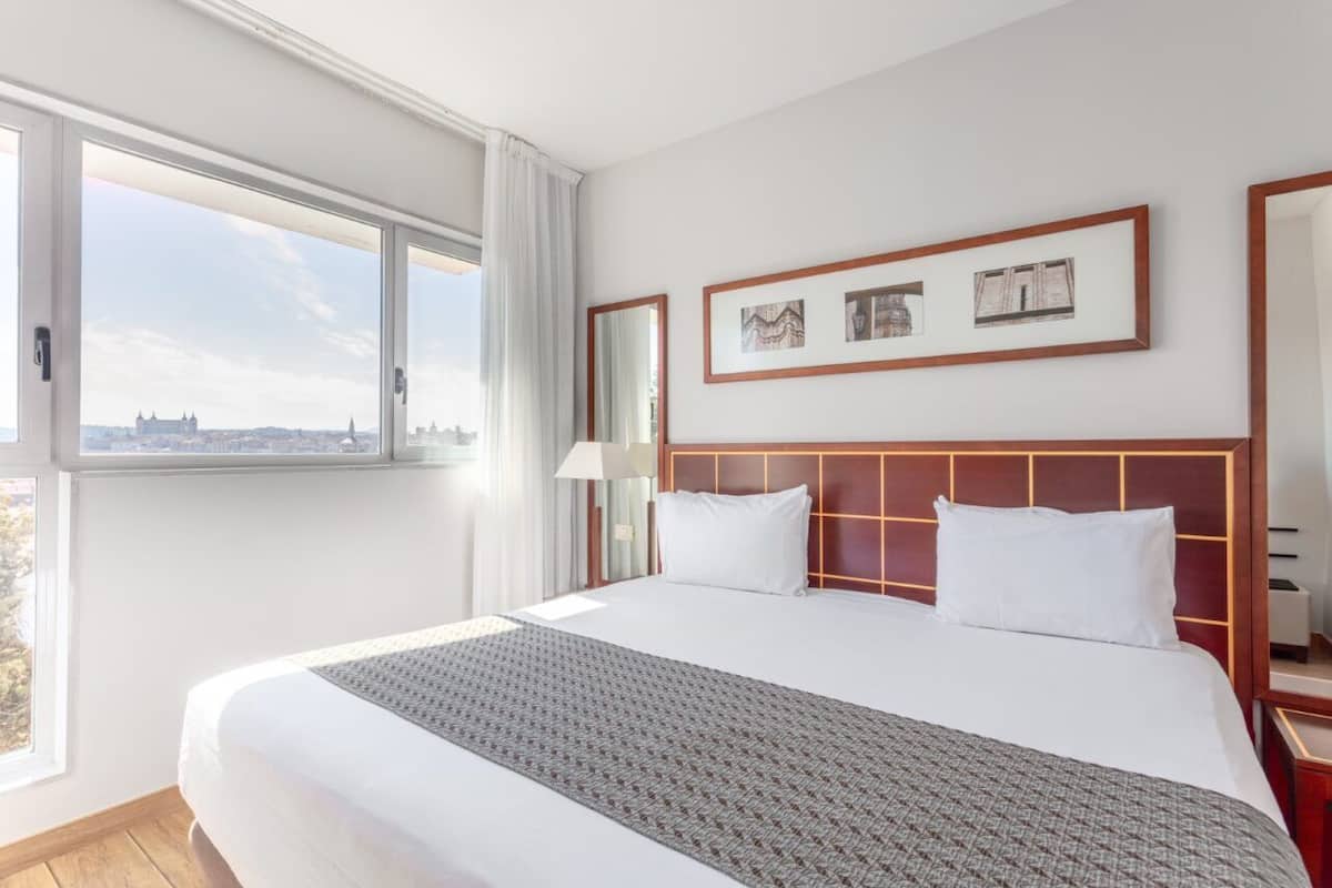 A hotel room with a large bed, a city view, and framed artwork on the wall.