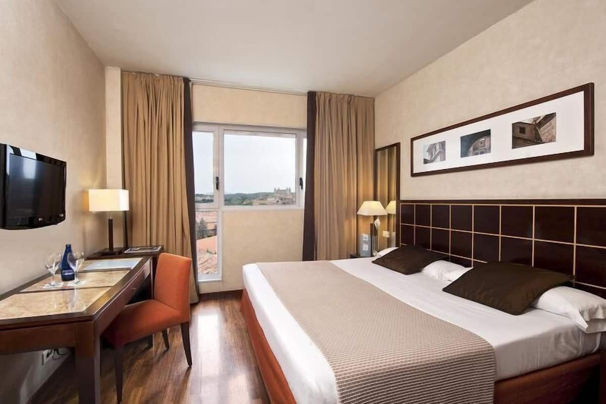 A hotel room with a bed, desk, chair, TV, and a window with curtains.