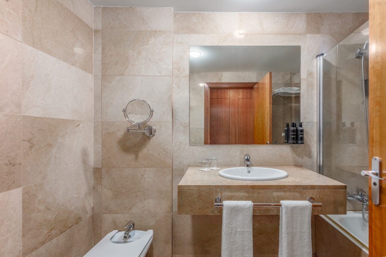 A modern bathroom with a marble-tiled wall, a wooden vanity, a white sink, a glass shower enclosure, and a mirror.