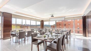 Un comedor moderno con amplias ventanas, varias mesas y sillas, y vistas a la ciudad.