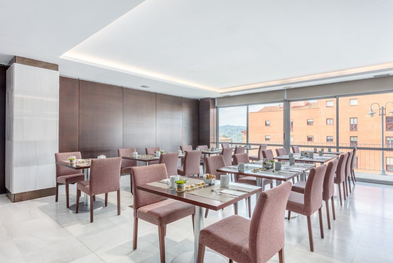 A modern dining area with multiple tables and chairs, large windows offering a city view, and a high ceiling with recessed lighting.
