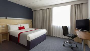 A hotel room with a bed, a desk with a TV, a chair, and a window with curtains.