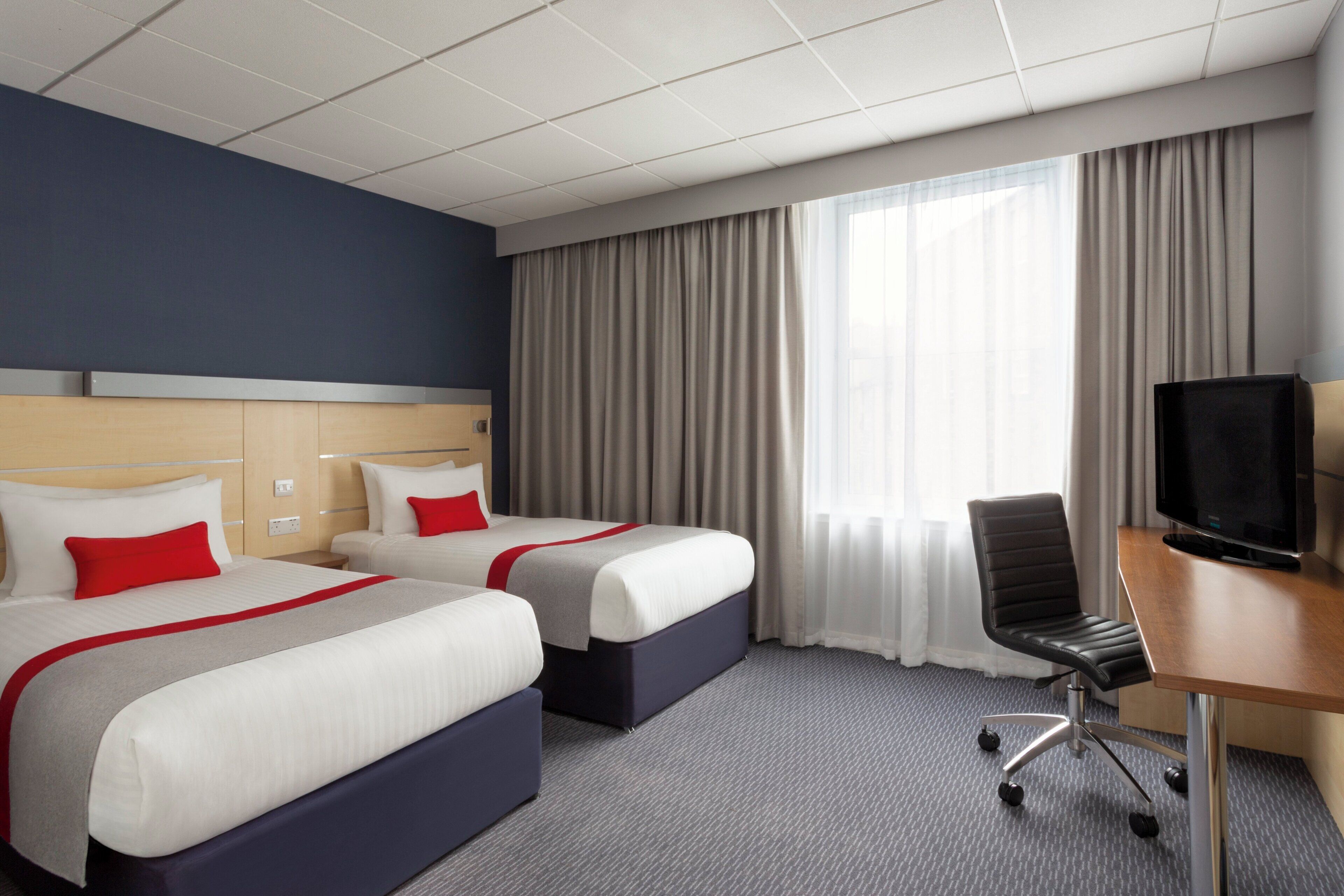 A hotel room with two beds, a desk with a chair, a television, and a large window with curtains.