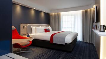 A hotel room with a bed, a desk, a chair, a TV, and a window with curtains.