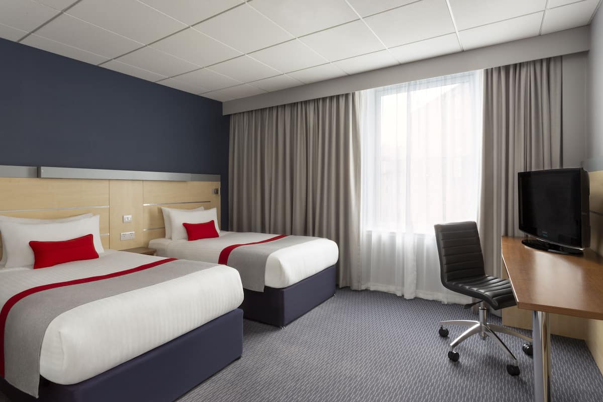 Ein Hotelzimmer mit zwei Betten, einem Schreibtisch mit Fernseher und einem großen Fenster mit Vorhängen.
