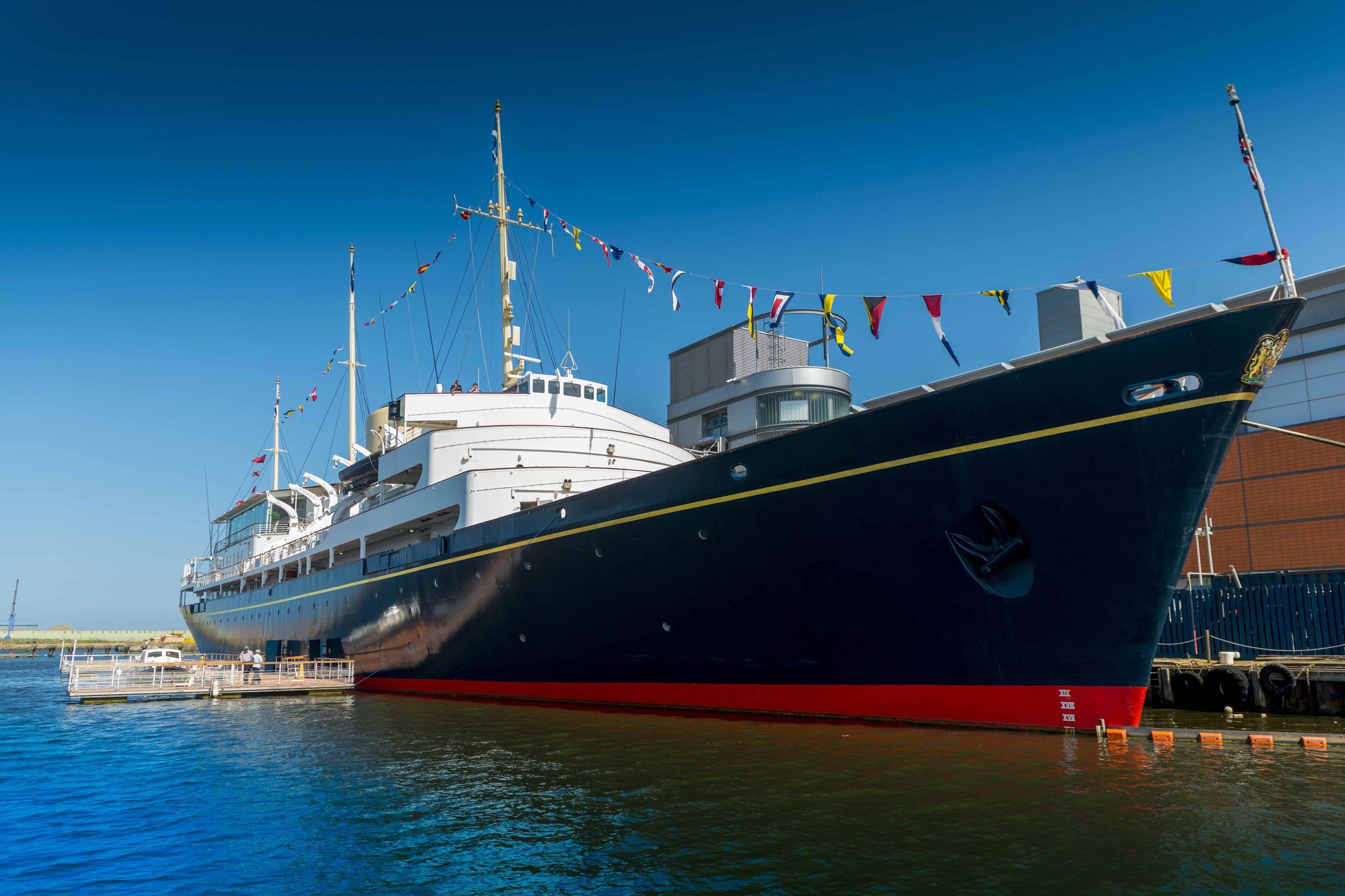 A large ship docked at a port with multiple flags, a cabin structure, and a red and black hull.