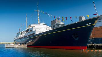 A large ship docked at a port with multiple flags, a cabin structure, and a red and black hull.